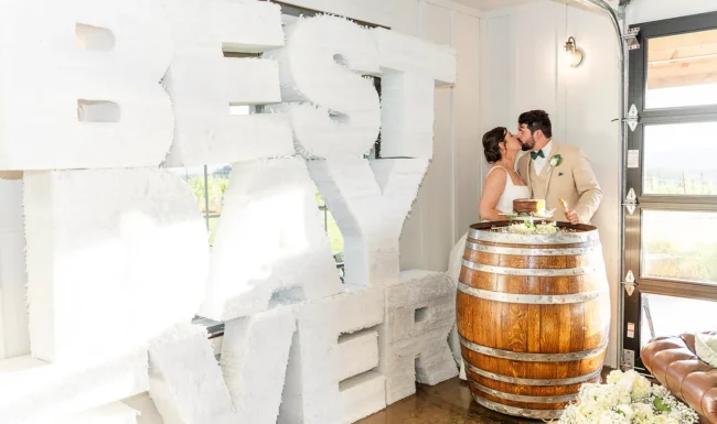 Bride and groom kissing after their cake cutting on a wine barrel with a Best Day Ever large sign